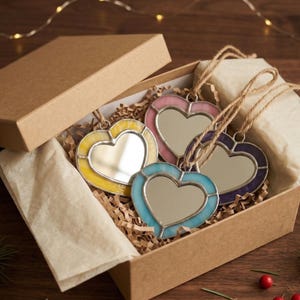 Puede incluir: Un conjunto de cuatro espejos en forma de corazón en una caja de regalo de cartón. Cada espejo tiene un borde de color amarillo, rosa, azul y morado. Los espejos están ensartados con hilo y descansan sobre papel triturado.