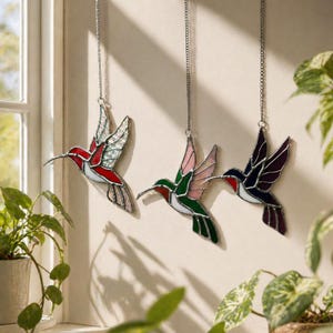 May include: Three stained glass hummingbird ornaments hanging from silver chains. The hummingbirds feature red, green, pink, and purple wings and bodies. The ornaments are suspended in front of a neutral wall, with a window and plants in the background.