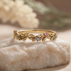 Puede incluir: Un anillo dorado con un diseño de hojas y pequeñas piedras preciosas de colores. El anillo presenta una banda adornada con hojas detalladas y está engastado con piedras preciosas verdes, amarillas, rosas, azules y turquesas. El anillo se muestra sobre una superficie de piedra blanca.