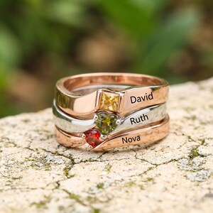 Puede incluir: Un conjunto de anillos apilables con tres bandas de acabados metálicos. La banda superior es de color rosa con una piedra cuadrada amarilla y el nombre David. La banda central es de color plateado con una piedra verde en forma de corazón y el nombre Ruth. La banda inferior es de color rosa con una piedra redonda roja y el nombre Nova.