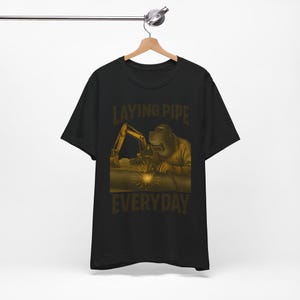 May include: Black t-shirt with a gold graphic of a welder working on a pipe, with the text "LAYING PIPE EVERYDAY". The design has a construction theme.