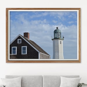 May include: A white lighthouse with a green roof stands tall against a blue sky with white clouds. A brown house with white windows is in the foreground. The image is framed in a wooden frame.