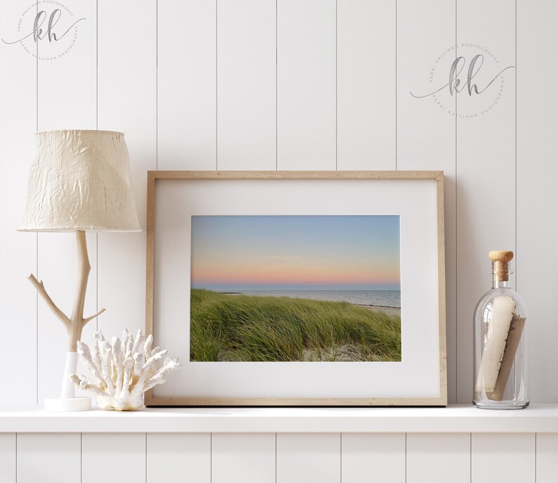 May include: A framed photograph of a sunset over a grassy beach. The sky is a gradient of pink, orange, and blue. The grass is green and blowing in the wind.