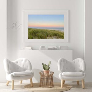 May include: A framed photograph of a beach scene with green grass in the foreground and a pink and blue sunset in the background. Two white chairs with wooden legs and a small wooden table with a potted cactus plant are in the foreground.