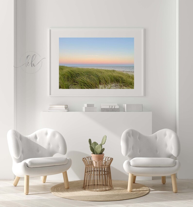 May include: A framed photograph of a beach scene with green grass in the foreground and a pink and blue sunset in the background. Two white chairs with wooden legs and a small wooden table with a potted cactus plant are in the foreground.