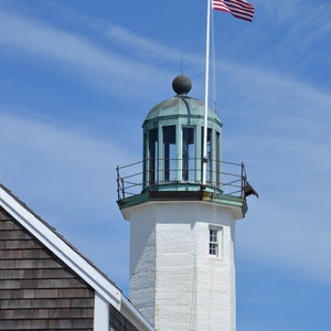 May include: A white lighthouse with a green roof and a white flagpole with an American flag flying from it. The lighthouse is against a blue sky with white clouds.