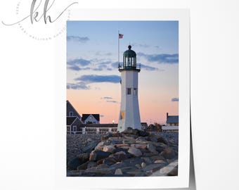 May include: A white lighthouse with a green roof stands on a rocky shore at sunset. The lighthouse is tall and slender, with a small American flag flying from its top. The sky is a soft pink and orange, and the water is calm and blue.