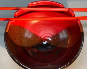May include: A ceramic bowl with a glossy red and brown glaze, designed to hold food. Two bright red chopsticks rest across the top of the bowl, creating a striking contrast. The bowl's interior has a dark, almost black center.