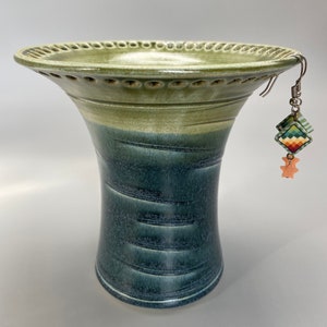 May include: A green and blue ceramic vase with a wide, flared top and a decorative rim. The vase has a textured, striped pattern and is decorated with a pair of colorful earrings.