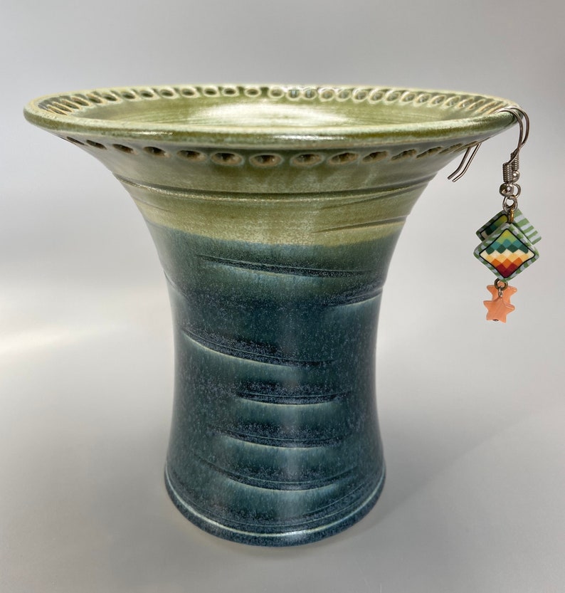 May include: A green and blue ceramic vase with a wide, flared top and a decorative rim. The vase has a textured, striped pattern and is decorated with a pair of colorful earrings.