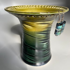 May include: A green ceramic vase with a textured surface and a wide, scalloped rim. The vase has a light green top and a dark green bottom. A pair of silver earrings with blue and green glass accents are hanging from the rim.