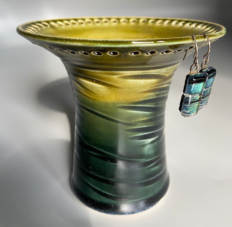 May include: A green ceramic vase with a textured surface and a wide, scalloped rim. The vase has a light green top and a dark green bottom. A pair of silver earrings with blue and green glass accents are hanging from the rim.