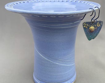 May include: A light blue ceramic vase with a wide, flared rim featuring a decorative perforated edge. A pair of triangular metal earrings with gold-toned celestial motifs, including a sun, moon, and stars, hangs from the rim of the vase.