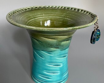 May include: A ceramic vase with a green and blue glaze. The vase has a wide, flared opening and a narrow base. The opening has a decorative edge and a small hole for hanging earrings.