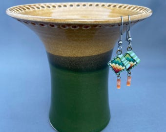 May include: A handcrafted ceramic vase with a wide, fluted rim and a green base. The vase has a layered color scheme of beige, brown, and green. A pair of earrings with square, patterned pendants and orange accents hang from the rim.