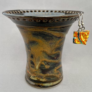 May include: A handcrafted ceramic vase with a flared top and a dark brown rim. The vase has a unique glaze with shades of brown, gold, and black. A pair of square, colorful earrings hangs from the vase, adding a pop of color.