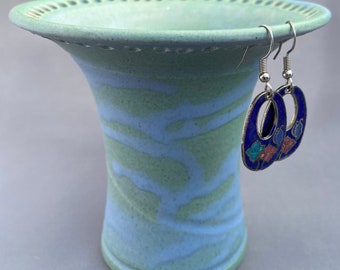 May include: A light blue ceramic vase with a scalloped rim and a swirling pattern. The vase has a pair of blue and silver earrings with floral accents hanging from the rim.