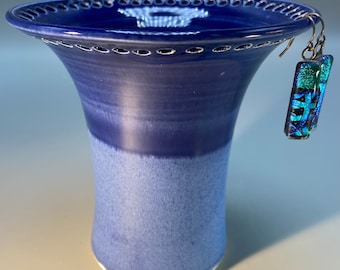 May include: A blue ceramic vase with a white rim and a decorative pattern around the top. The vase has a gradient of blue from dark to light. A pair of blue and green glass earrings are hanging from the vase.