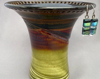 May include: A handcrafted ceramic vase with a flared rim and a gradient of colors, transitioning from green at the base to brown and black at the top. Two dichroic glass earrings hang from the rim, reflecting blue and green hues.