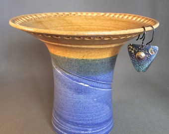 May include: A handcrafted ceramic vase with a wide, fluted rim and a flared base. The vase features a color gradient of tan, blue, and purple. A pair of triangular earrings with a celestial design hangs from the rim, featuring sun, moon, and star motifs.