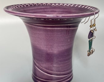 May include: A purple ceramic vase with a decorative rim and a swirling design. The vase has a small hook on the rim for hanging earrings.