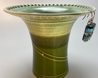 May include: A handcrafted, green ceramic vase with a flared rim and decorative perforations. The vase has a gradient of green hues and features two curved, etched lines. A pair of dichroic glass earrings hangs from the rim.