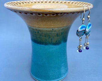 May include: A handcrafted ceramic vase with a flared rim and a turquoise base, transitioning to a brown top. Two dangle earrings with blue and white marbled oval beads and small purple and white beads hang from the rim.