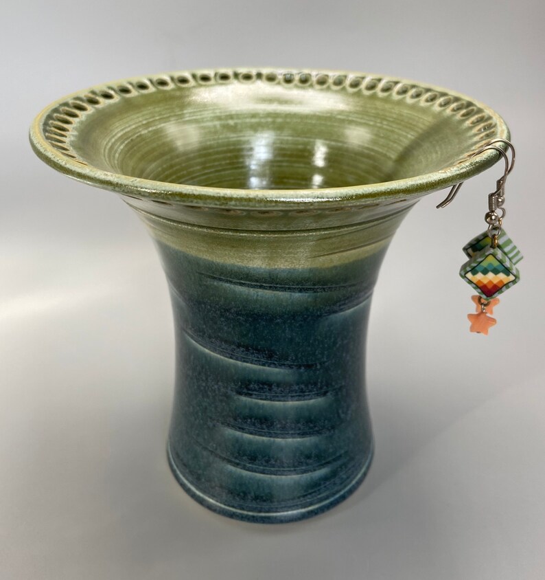 May include: A green and blue ceramic vase with a decorative rim and a pair of earrings hanging from the rim.