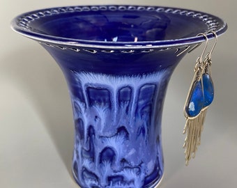 May include: A cobalt blue ceramic vase with a wide, decorative rim and a flared shape. The vase has a textured, dripped glaze effect in shades of blue and white. A pair of silver earrings with blue gemstone pendants and fringe hang from the rim.