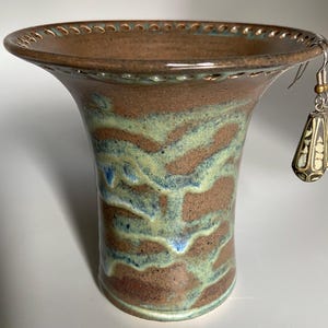 May include: A brown ceramic vase with a green and blue glaze. The vase has a scalloped rim and a decorative earring hanging from it.