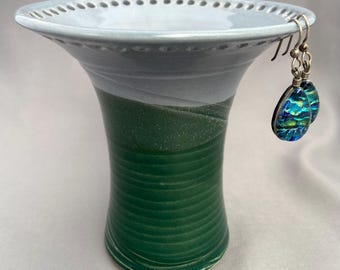 May include: A ceramic bowl with a wide, flared rim featuring a dotted pattern, glazed in light gray at the top and dark green with horizontal ridges at the base. A pair of teardrop-shaped dichroic glass earrings with blue and green hues hang from the rim.