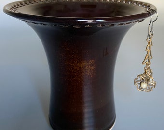May include: A dark brown ceramic vase with a decorative rim and a white base. An earring with a gold-colored design hangs from the rim of the vase.