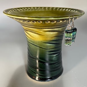 May include: A green ceramic vase with a wide, scalloped rim and a textured, ombre finish. The vase has a dark green base and fades to a lighter green at the top. A pair of silver earrings with green and gold glass accents are hanging from the rim.