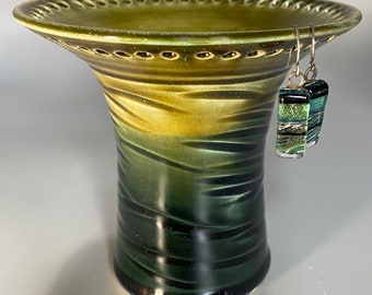 May include: A green ceramic vase with a wide, scalloped rim and a textured, ombre finish. The vase has a dark green base and fades to a lighter green at the top. A pair of silver earrings with green and gold glass accents are hanging from the rim.