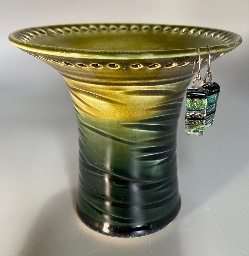 May include: A green ceramic vase with a wide, scalloped rim and a textured, ombre finish. The vase has a dark green base and fades to a lighter green at the top. A pair of silver earrings with green and gold glass accents are hanging from the rim.