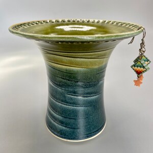 May include: A green and blue ceramic vase with a wide, flared top and a narrow base. The vase has a textured, speckled glaze and a decorative rim. A pair of colorful earrings with a star charm hang from the rim of the vase.