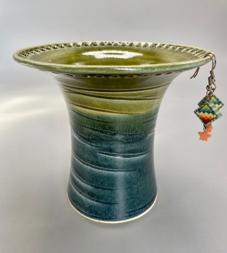 May include: A green and blue ceramic vase with a wide, flared top and a narrow base. The vase has a textured, speckled glaze and a decorative rim. A pair of colorful earrings with a star charm hang from the rim of the vase.