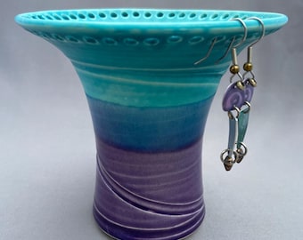 May include: A ceramic vessel with a wide, perforated rim, featuring a gradient glaze that transitions from turquoise at the top to deep blue and purple at the base. A pair of dangling earrings with purple and teal metal components hangs from the rim.