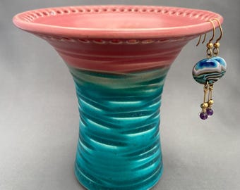 May include: A ceramic bowl with a wide, flared rim featuring a pink interior and a textured, wavy teal exterior. A pair of dangling earrings hangs from the rim, showcasing blue, white, and brown swirled glass beads with small purple accents.