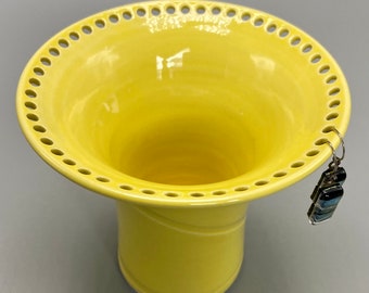 May include: A yellow ceramic vase with a scalloped rim and a decorative hole pattern. The vase has a flared top and a narrow base.