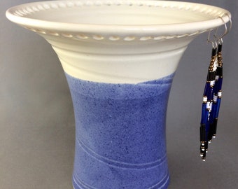 May include: A white ceramic vase with a blue glaze and a scalloped rim. The vase has a beaded design around the top edge. A pair of blue and white beaded earrings are hanging from the rim.