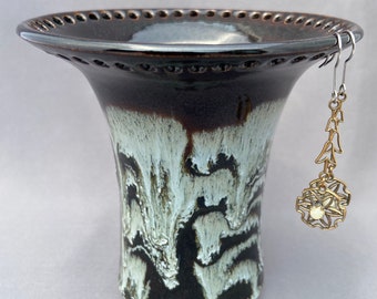 May include: A brown and white ceramic vase with a scalloped rim and a decorative design. A pair of gold earrings with a floral design hangs from the rim of the vase.