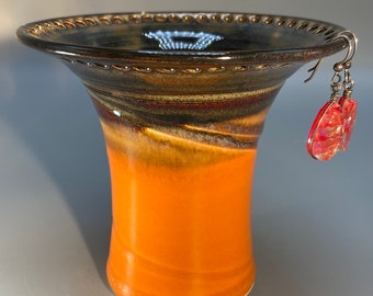 May include: A handmade ceramic vase with a flared rim, featuring a gradient of colors from orange to brown and black. The vase has a textured surface and is paired with red, translucent earrings.