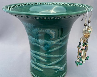 May include: A green ceramic vase with a wide, scalloped rim and a decorative design of blue and white lines. A pair of dangling earrings with beads and charms are hanging from the rim.