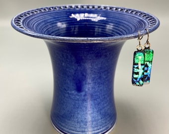 May include: A cobalt blue vase with a flared top and a decorative rim. A pair of rectangular earrings with green and blue dichroic glass hang from the vase. The earrings have gold-colored ear wires.