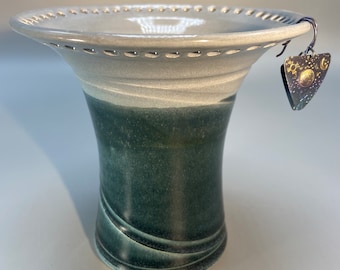 May include: A white and green ceramic vase with a wide, scalloped rim and a decorative hole pattern. The vase has a dark green glaze with a light green swirl pattern. A small, triangular metal earring with a sun and moon design hangs from the rim.