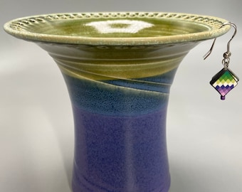 May include: A ceramic vase with a green, yellow, blue, and purple glaze. The vase has a wide mouth and a narrow base. It has a decorative rim and a hook on the side.