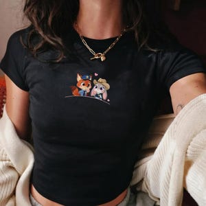 May include: Black crop top featuring an embroidered design of two cartoon characters, a fox in a police uniform and a bunny in a cowboy hat, with pink hearts above them. The shirt has short sleeves and a crew neck.