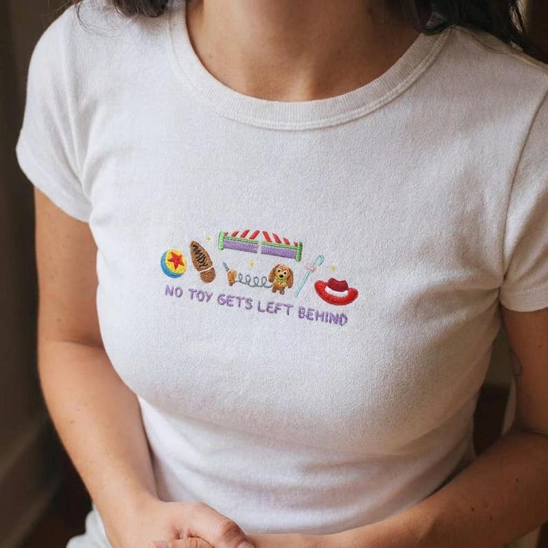 Pu&ograve; includere: Una t-shirt bianca a maniche corte con un disegno ricamato di vari giocattoli colorati e il testo "NO TOY GETS LEFT BEHIND" in lettere viola. Il ricamo include una palla, uno stivale, un cane a molla e un cappello da cowboy rosso.