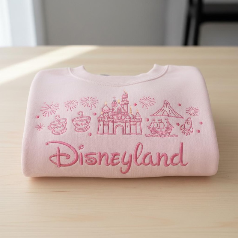 May include: A light pink sweatshirt with a Disneyland design. The design includes a castle, teacups, a volcano, a ship, and a rocket, all in pink. The word "Disneyland" is written in a cursive font.